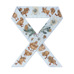 A woman in a beige top and skirt pairs her look with the SABINA SAVAGE LONDON BRIDGE IS FALLING DOWN SILK RIBBON by SABINA SAVAGE ENGLAND, elegantly tied around her neck against a gray backdrop, with her hands clasped in front.