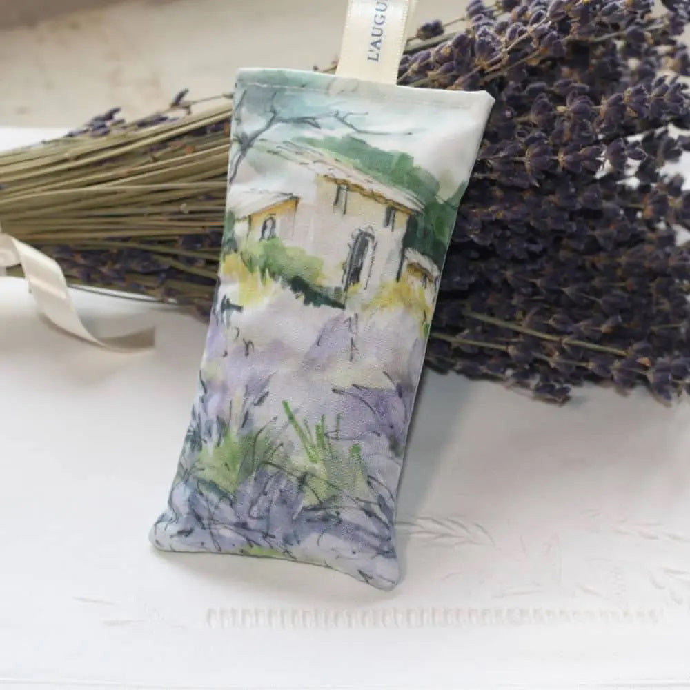 The L&#39;AUGUSTE PROVENCE - FRENCH LAVENDER SACHET FROM PROVENCE features a painted house in a lavender field, stands upright with dried lavender in front, and is topped with a white &quot;L&#39;AUGUSTE&quot; ribbon—authentic from Provence.