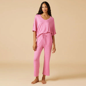 A woman stands barefoot facing away, wearing the SOFTIES Dream Relaxed V-Neck with Capri Lounge Set in Fuschia Stripe—soft jersey pink and white striped pajamas with a loose top and wide-leg pants—against a plain beige background.