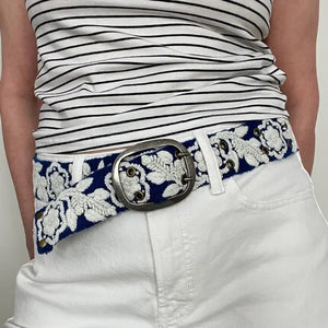 The JENNY KRAUSS Two Toned Belted in Blue and Cream features intricate embroidery, a silver rectangular buckle, and is handcrafted in Peru. Shown coiled on a white surface.
