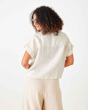 A woman with curly hair wears sunglasses and the MERSEA HADLEY BOXY LINEN SHIRT in cream, featuring loose short sleeves, paired with beige high-waisted pants with front pockets, standing against a white background.