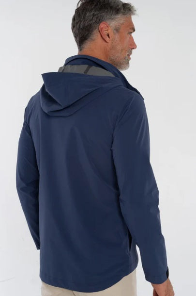 A man wearing the CRIQUET - Watershed Rain Jacket in Navy stands outdoors on a rainy day, with trees and wet pavement in the background.