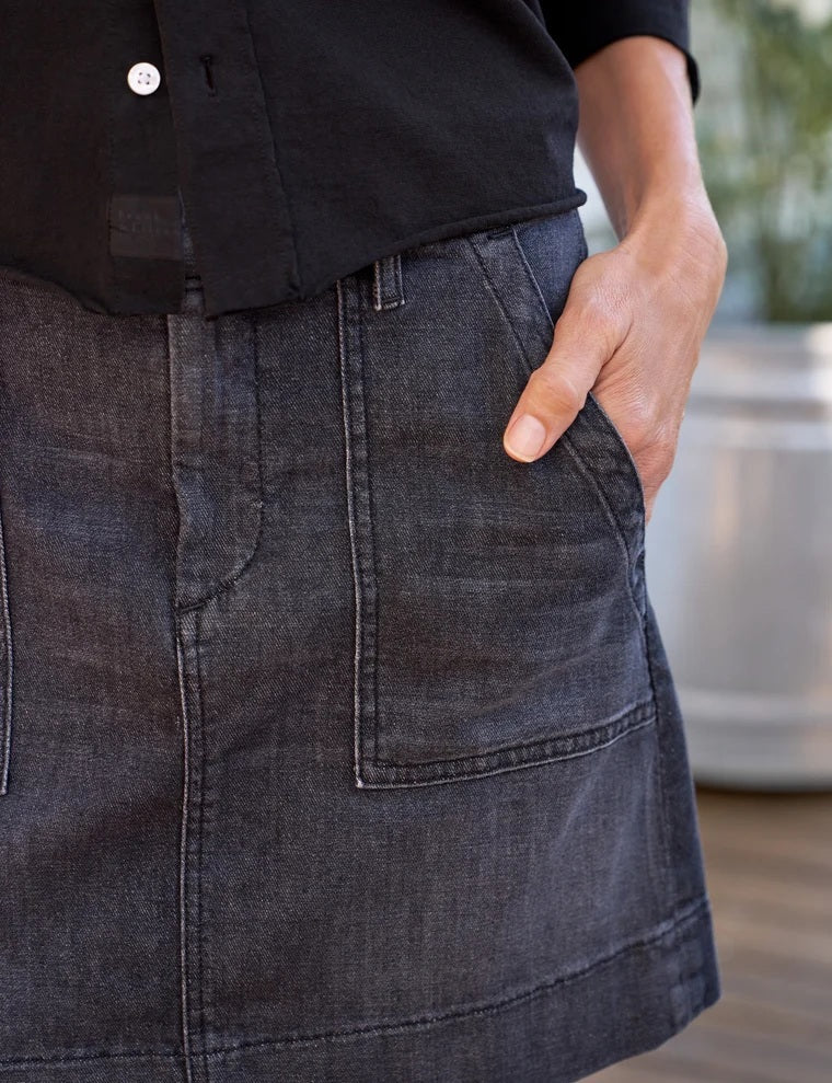 A person wearing a black button-up shirt and the FRANK & EILEEN Jameson Utility Skirt in Italian Dream Light Denim 1981 Wash stands with one hand in a pocket. The background is blurred with some greenery visible.