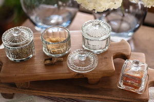 The ETUHOME Bistro Glass Sugar Cellar with Scoop by ETUHOME holds brown sugar on a white countertop near a folded white towel with red stripes and a wooden tray, adding classic charm to your kitchen storage collection.