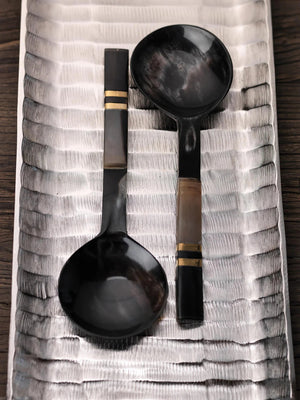 The SEYCHELLES HORN SALAD SERVER SET by ZODAX features two dark-hued serving spoons with round bowls and gold-tan accented handles, ideal for luxury home décor or as an elegant hostess gift. Displayed on a white background.