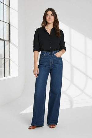 A person models the SPANX - SPANXSHAPE AUTHENTIC 360 WIDE LEG JEAN by SPANX, featuring a high-waisted fit and crafted from 4-way stretch denim, paired with a black shirt. Only the lower half of their body is visible as they stand with one hand at their side against a plain background.