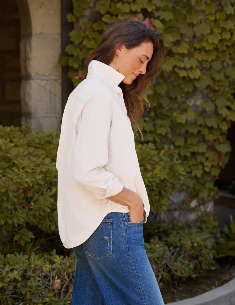 A woman stands indoors near a window, wearing the FRANK &amp; EILEEN Eileen Relaxed Button Up Shirt in Italian Corduroy Vintage White, paired with blue jeans and brown sandals. She has one hand in her pocket and is smiling softly.