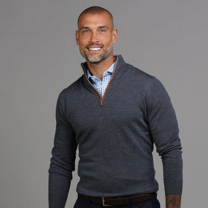 A smiling man with a shaved head and short beard wears the COLLARS AND CO Charcoal with Brown Suede Trim Cashmere Touch Australian Merino Quarter Zip Sweater over a blue checkered shirt, standing against a plain gray background.