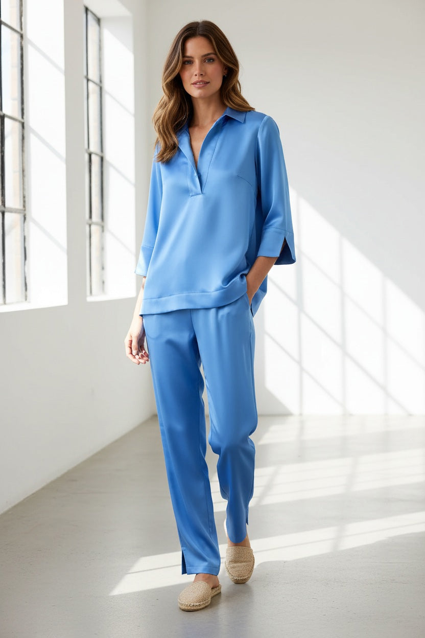 A woman with blonde hair wears the HINSON WU Aileen 3/4 Sleeve Liquid Satin Shirt in Sky Blue—a loose-fitting blouse featuring an elegant drape and luminous sheen. Paired with matching pants, the light blue satin fabric from HINSON WU catches the light beautifully as she stands against a simple, light background.