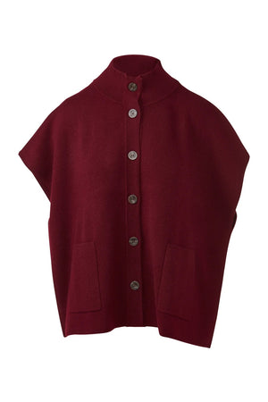 A woman stands indoors by a window, smiling softly as she wears the KINROSS CASHMERE Short Sleeve Button Mock Cardigan in a rich burgundy hue over a black top, blue jeans, and brown ankle boots. Her long hair falls freely around her shoulders.