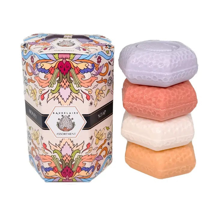 Four pastel-colored, triple-milled soaps—lavender, pink, white, and orange—are stacked next to a decorative floral-patterned box labeled “BAUDELAIRE - HONEY SOAP ASSORTMENT 1.4 OZ QUARTET” by BAUDELAIRE.