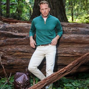 A man in white pants stands before a large fallen tree in the forest, wearing the COLLARS AND CO Hunter Green with Brown Suede Trim Cashmere Touch Australian Merino Wool Sweater, with one hand in his pocket and a brown leather backpack by his side.