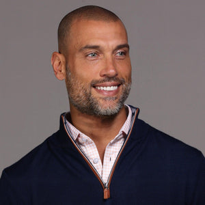 A man with short hair and a beard stands against a gray background wearing the COLLARS AND CO Navy with Brown Suede Trim Cashmere Touch Australian Merino Quarter Zip Sweater over a white shirt, khaki pants, and brown high-top sneakers, one hand in his pocket.