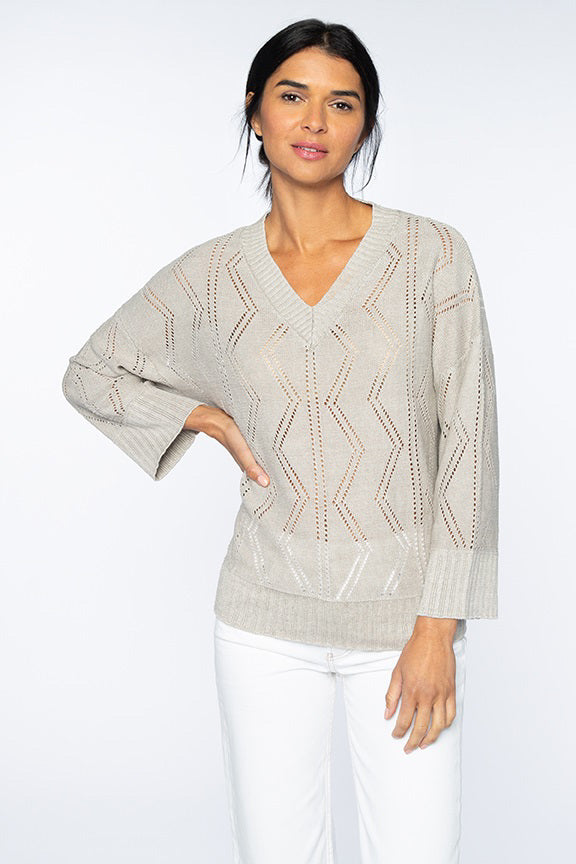 A person with long dark hair is wearing a KINROSS CASHMERE OPENWORK VEE NECK SWEATER by KINROSS, featuring a light gray knit made of 100% linen and a chevron pattern. They have paired it with white pants, standing against a plain white background with one hand resting on their hip.
