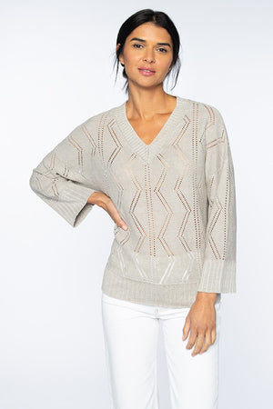 A person with long dark hair is wearing a KINROSS CASHMERE OPENWORK VEE NECK SWEATER by KINROSS, featuring a light gray knit made of 100% linen and a chevron pattern. They have paired it with white pants, standing against a plain white background with one hand resting on their hip.