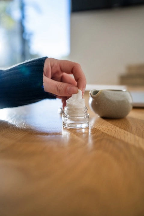 A hand pours liquid from a small ceramic pitcher into a glass jar filled with a white, fluffy substance atop a wooden surface, demonstrating the preparation of essential oil tablets for use with the MADISON AND GREEN - REST YOUR EYES AT THE DESK aromatherapy eye covers or reusable eye mask by MADISON GREEN.