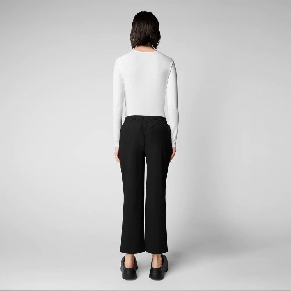 A person with shoulder-length hair and a neutral expression wears a white long-sleeve shirt, SAVE THE DUCK MILAN PULL ON PANT in black wrinkle-free fabric, and black slip-on shoes. Their hands rest at their sides against a plain background.
