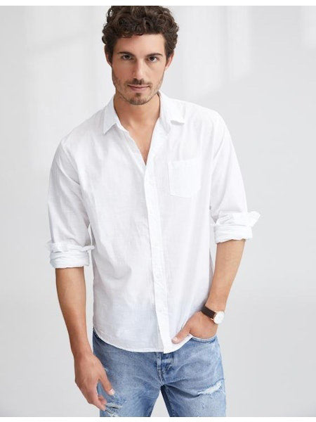 A man with short curly hair wears the FRANK &amp; EILEEN LUKE Men's Button Up Shirt in Classic White Poplin (sleeves rolled), paired with light blue ripped jeans and a watch, smiling slightly against a plain background.