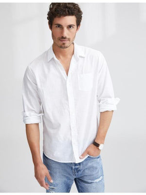 A man with short curly hair wears the FRANK & EILEEN LUKE Men's Button Up Shirt in Classic White Poplin (sleeves rolled), paired with light blue ripped jeans and a watch, smiling slightly against a plain background.