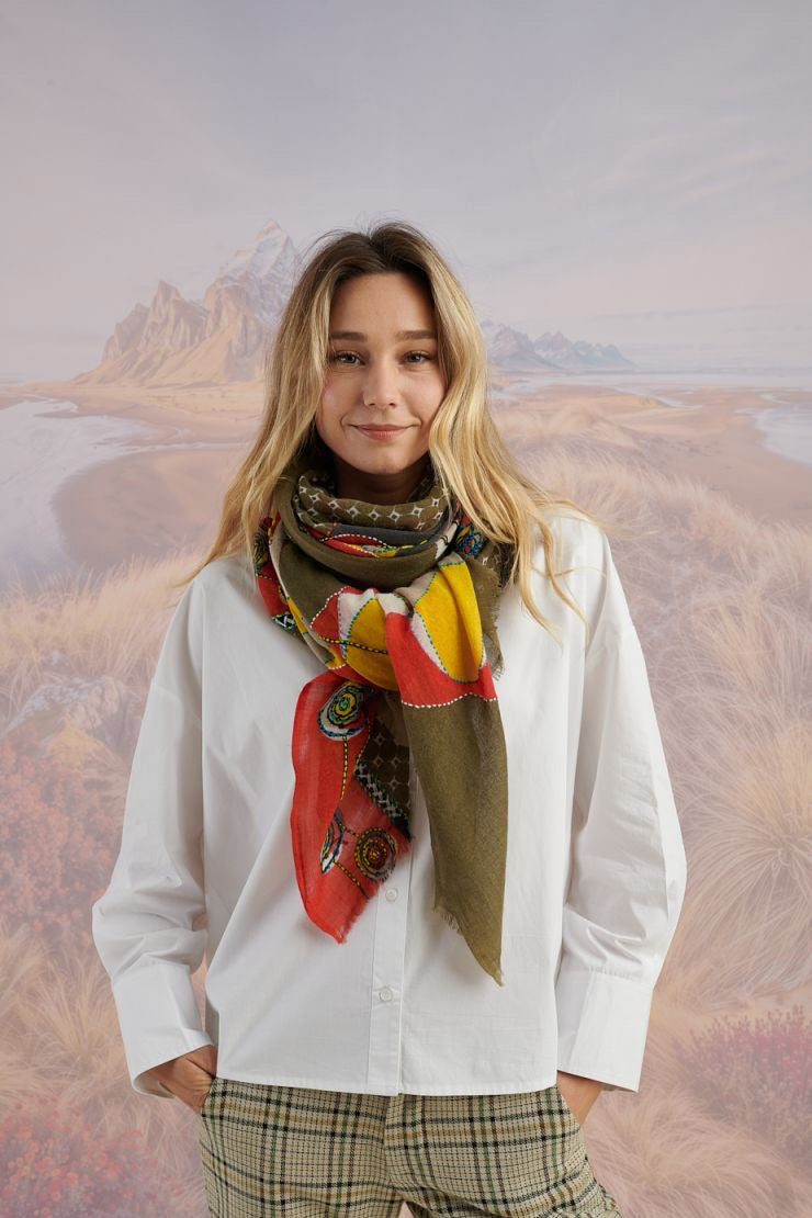 A woman with long blonde hair, in a light blue shirt and dark pants, smiles in front of a scenic backdrop, draping the elegant, hand-embroidered STORIATIPIC - ASTER WOOL SCARF around her shoulders.