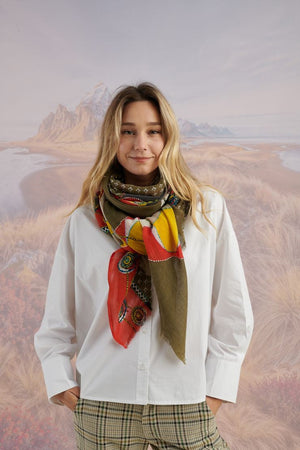 A woman with long blonde hair, in a light blue shirt and dark pants, smiles in front of a scenic backdrop, draping the elegant, hand-embroidered STORIATIPIC - ASTER WOOL SCARF around her shoulders.