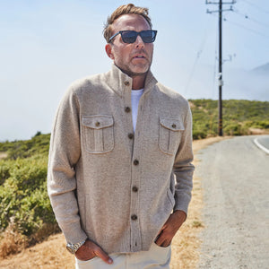 A man with short hair and a trimmed beard stands outdoors, wearing the COLLARS AND CO - NAPOLI CARDIGAN IN WOOL AND CASHMERE CAMEL over a white shirt, looking seriously at the camera with misty hills and green bushes in the background.