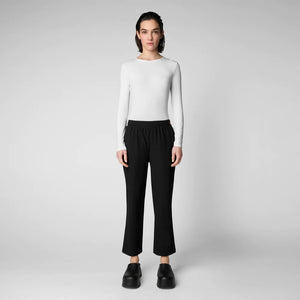 A person with shoulder-length hair and a neutral expression wears a white long-sleeve shirt, SAVE THE DUCK MILAN PULL ON PANT in black wrinkle-free fabric, and black slip-on shoes. Their hands rest at their sides against a plain background.