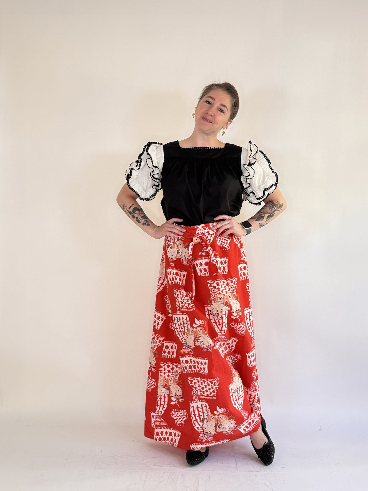 A person with tattooed arms stands against a light background, wearing a black top with sheer ruffled sleeves, black shoes, and the FARYLINA - FIFI WRAP SKIRT by FARYLINA featuring a white and beige cat pattern.