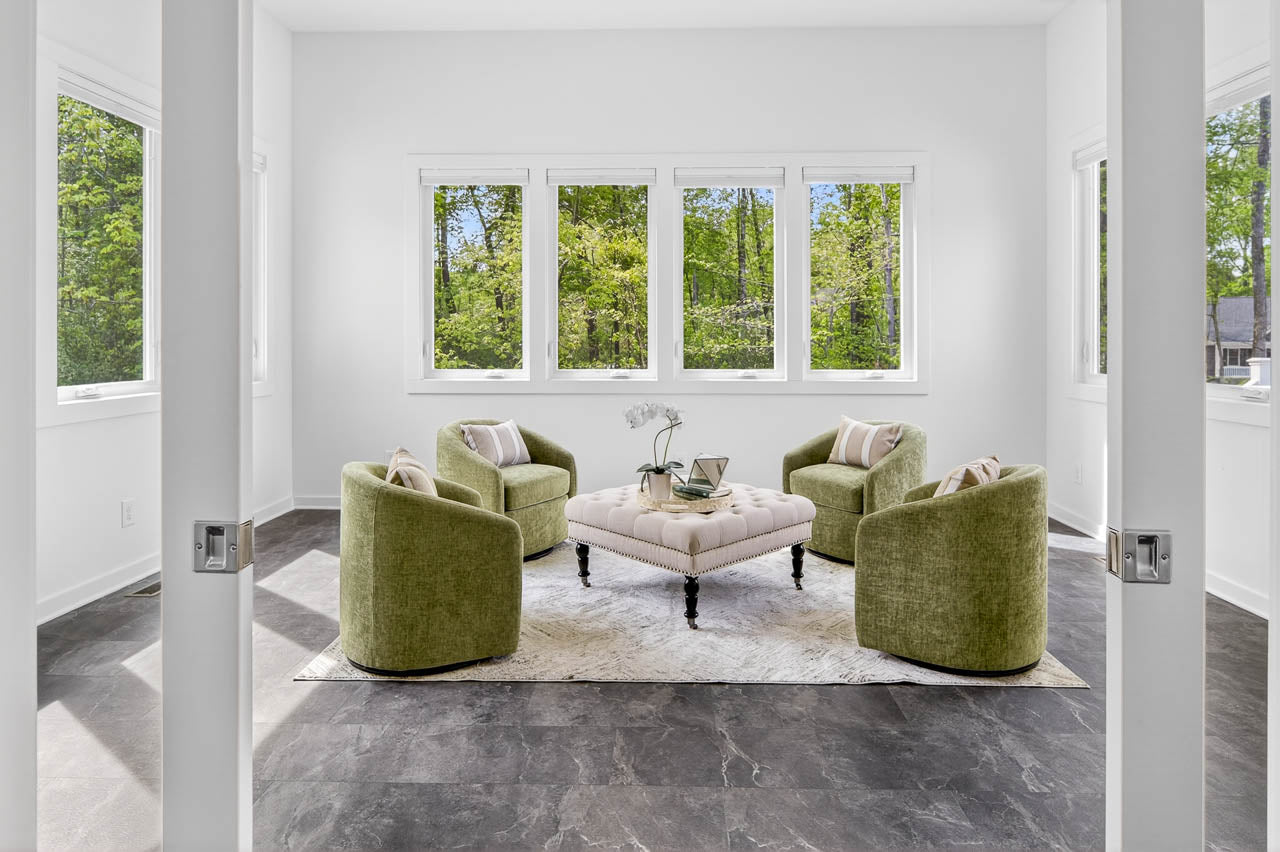 A bright, modern sitting room with four green armchairs arranged around a tufted beige ottoman on a white rug. Large windows offer views of lush trees outside. The space has white walls and dark tiled flooring.
