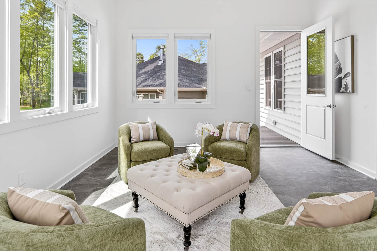 Bright sunroom with three green armchairs around a beige tufted ottoman, decorative tray and flowers on top. Large windows and an open door let in natural light; view of trees and another building outside.