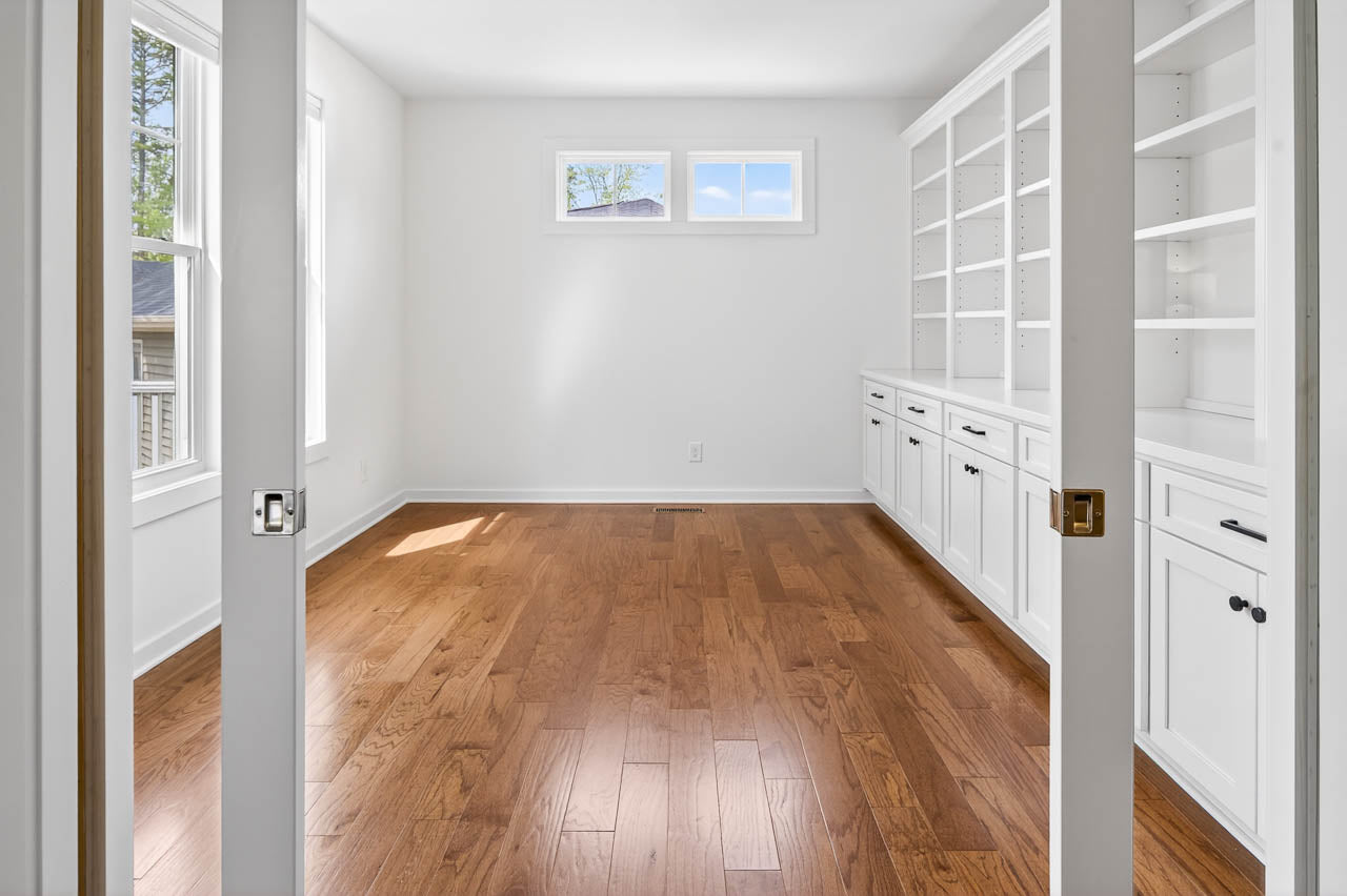 A bright, empty room with hardwood floors, white walls, built-in white shelves and cabinets on one side, and two large windows letting in natural light. Double glass doors open into the room.
