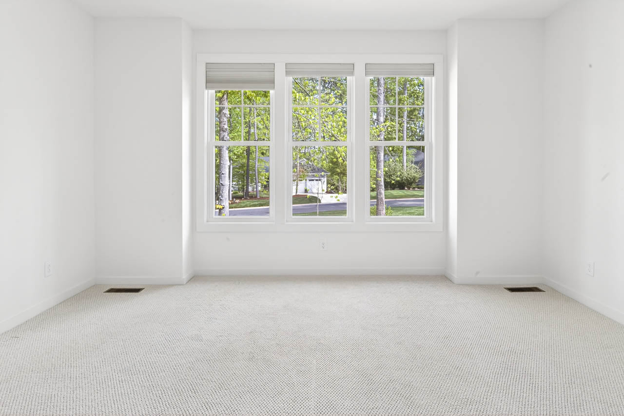 Bright, empty room with white walls and beige carpet, featuring a large three-pane window showing green trees and a suburban street outside.