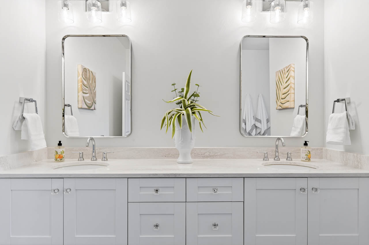 A modern bathroom with a double sink vanity, white cabinets, two rectangular mirrors, wall-mounted lights, and a white vase with green leaves between the sinks. Towels hang on both sides, and artwork is on the wall.