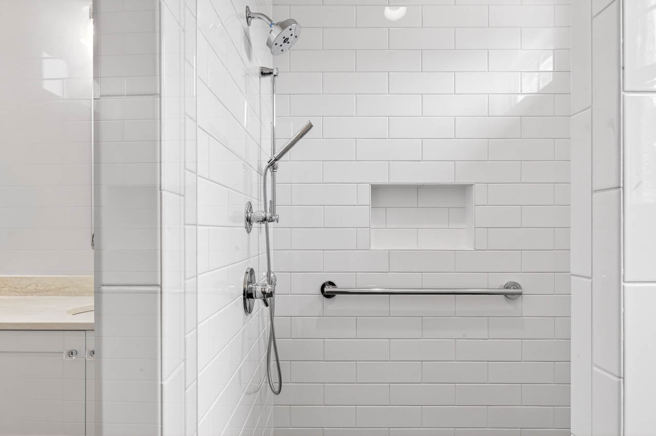 A modern shower with white subway tile, a wall-mounted showerhead, a handheld shower, a built-in shelf, and a horizontal grab bar for accessibility. Part of a vanity is visible on the left.