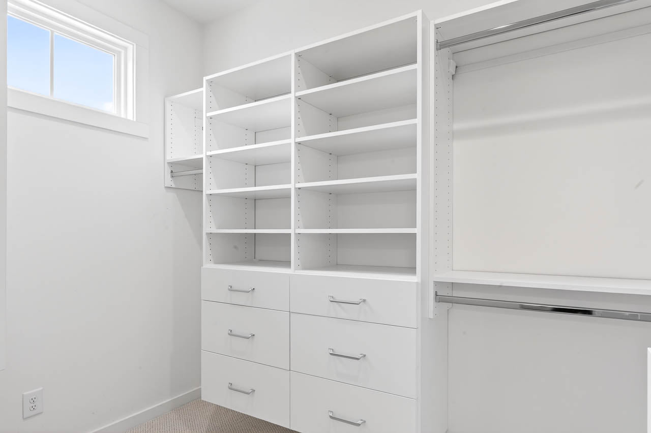 A bright, empty walk-in closet with white shelves, drawers, and hanging rods. There is a small window letting in natural light, and the walls and carpet are neutral in color.
