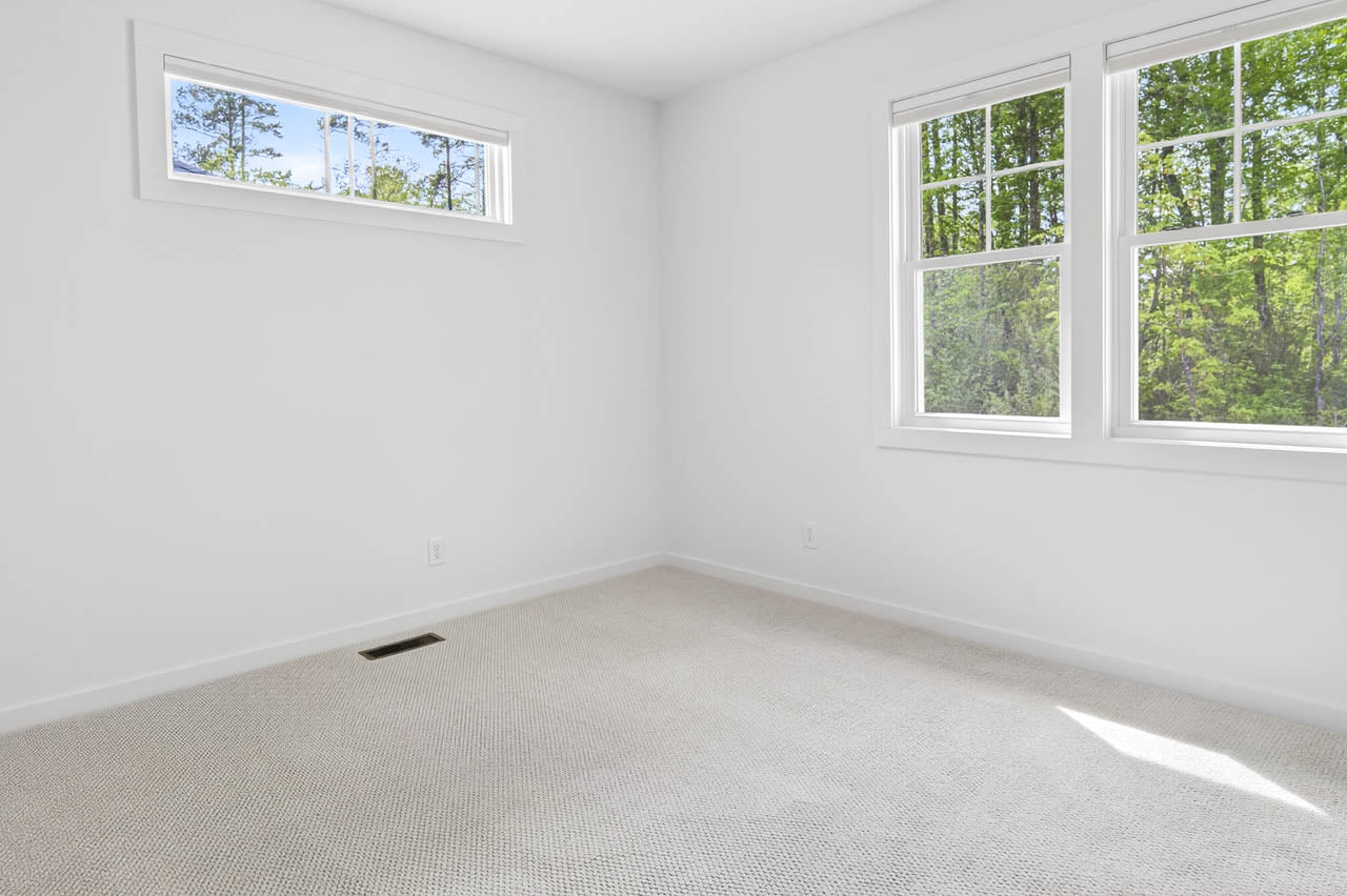A bright, empty room with white walls, beige carpet, and two large windows and one small window letting in natural light with a view of green trees outside.