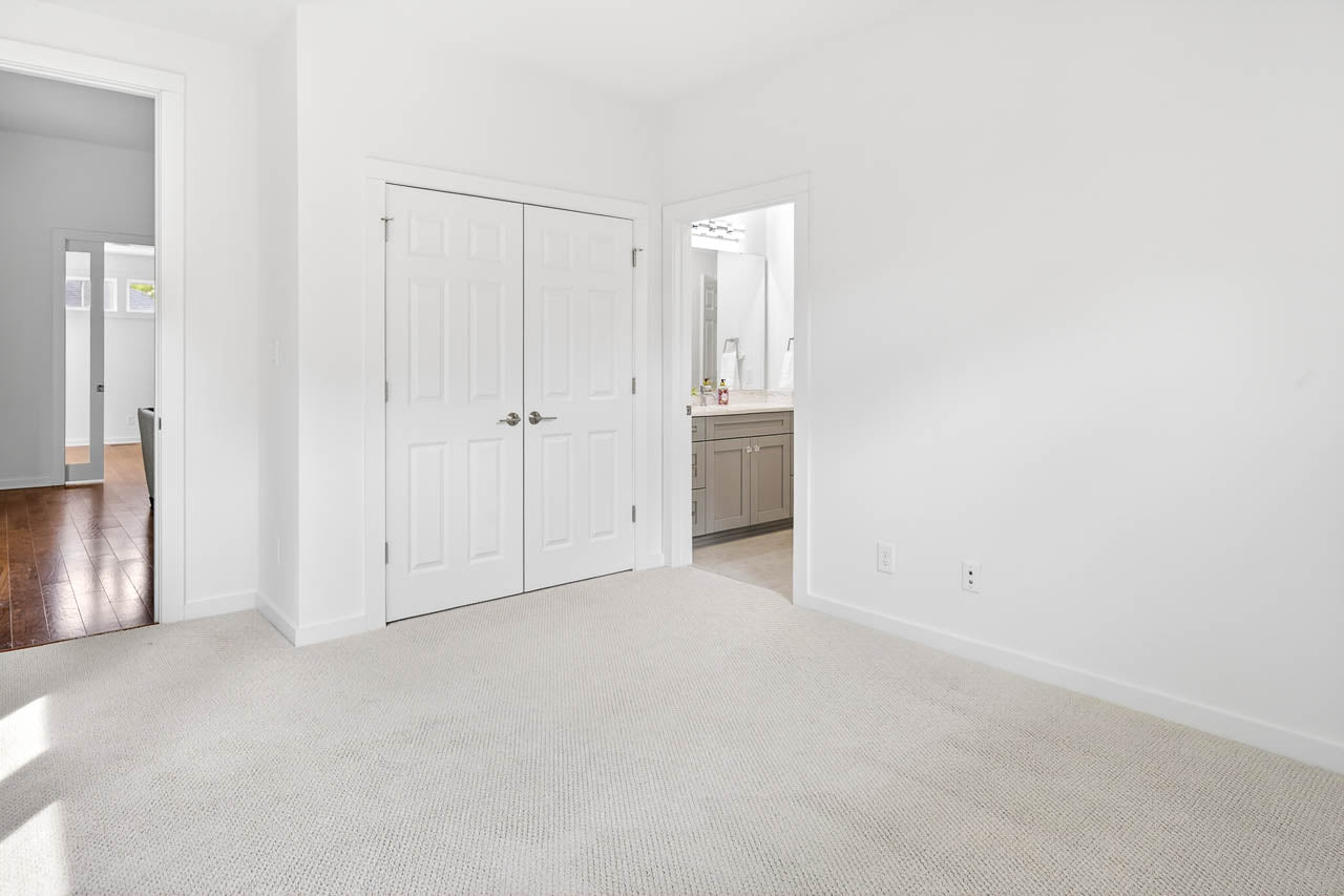 Bright, empty bedroom with white walls, light carpet, double-door closet, and an open doorway leading to a bathroom with a sink and mirror. Natural light enters from a window off-screen.