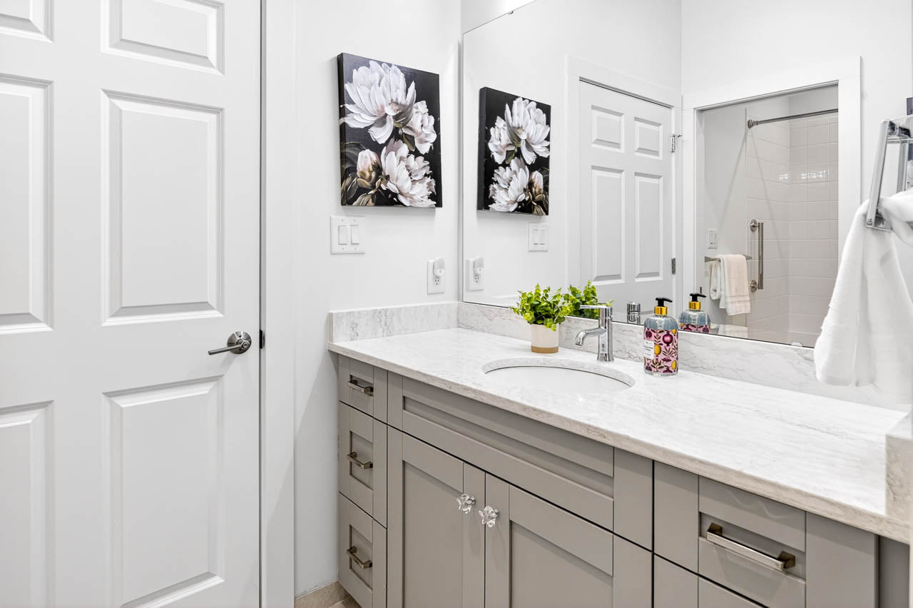 Modern bathroom with a white marble countertop, gray cabinets, a sink, potted plant, and soap dispensers. Two floral paintings hang above the vanity. A shower with a white curtain is visible in the reflection of the mirror.