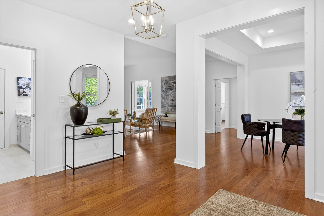 Bright, modern home interior with white walls, wood floors, and open layout. Entryway features a console table with vase and mirror, leading to a dining area, sunroom, and a bathroom on the left.