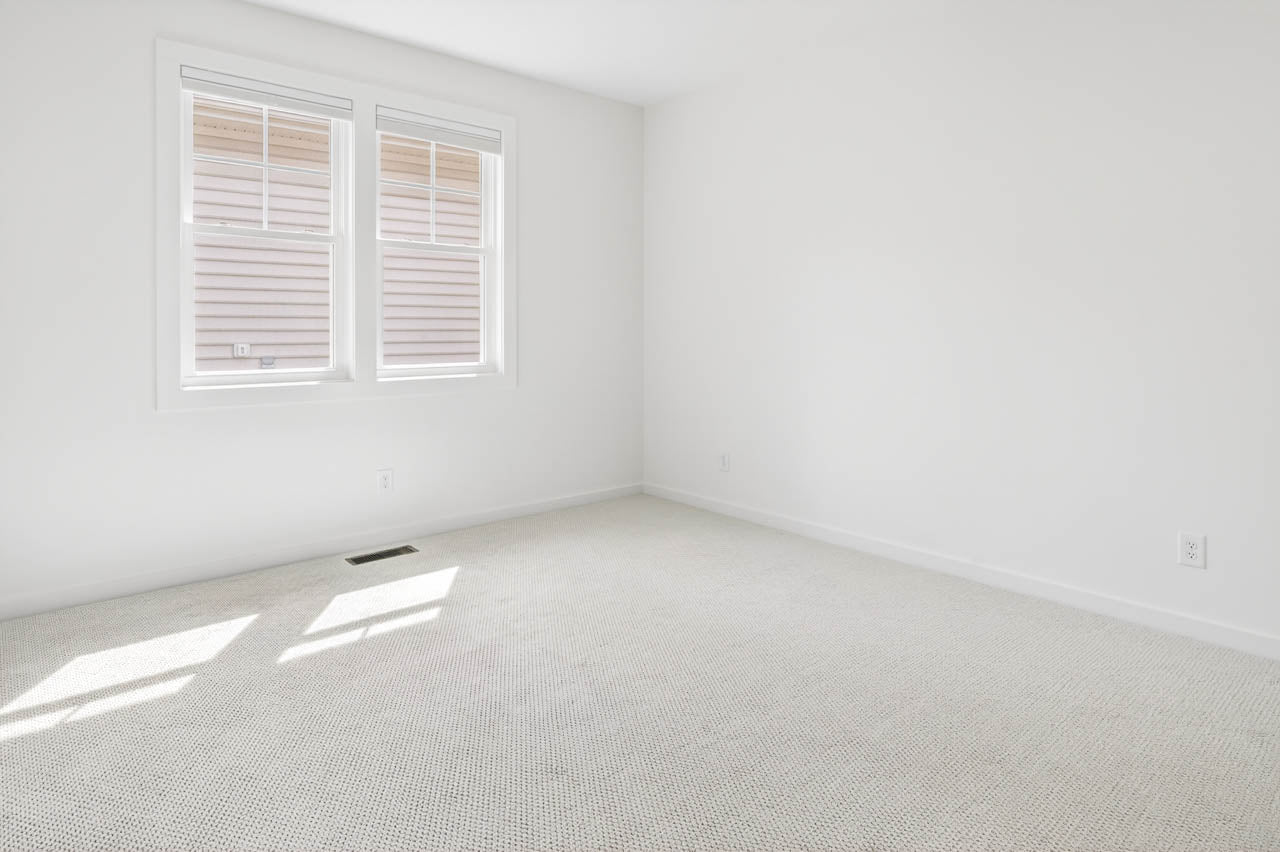 Empty, bright room with white walls, light-colored carpet, and two windows letting in natural light. The windows face an exterior wall of another building. The room has minimal features and a clean, modern look.