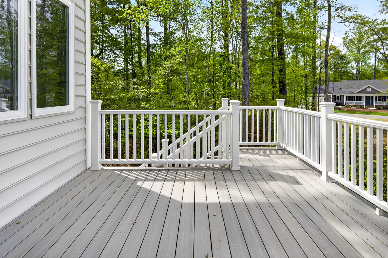 A spacious, light gray deck with white railing overlooks a wooded area and a nearby house. The deck is attached to a white-paneled home and features stairs leading down to the yard.