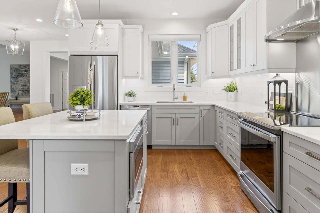 Modern kitchen with white cabinets, stainless steel appliances, marble countertops, and a central island with barstools. Hardwood floors and pendant lighting add warmth; a window above the sink brings in natural light.