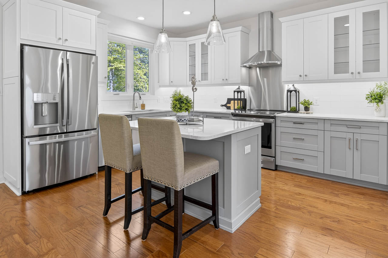 Modern kitchen with white cabinets, stainless steel appliances, a large refrigerator, a stove with a hood, an island with two upholstered chairs, pendant lights, and hardwood floors. Green plants add a fresh touch.
