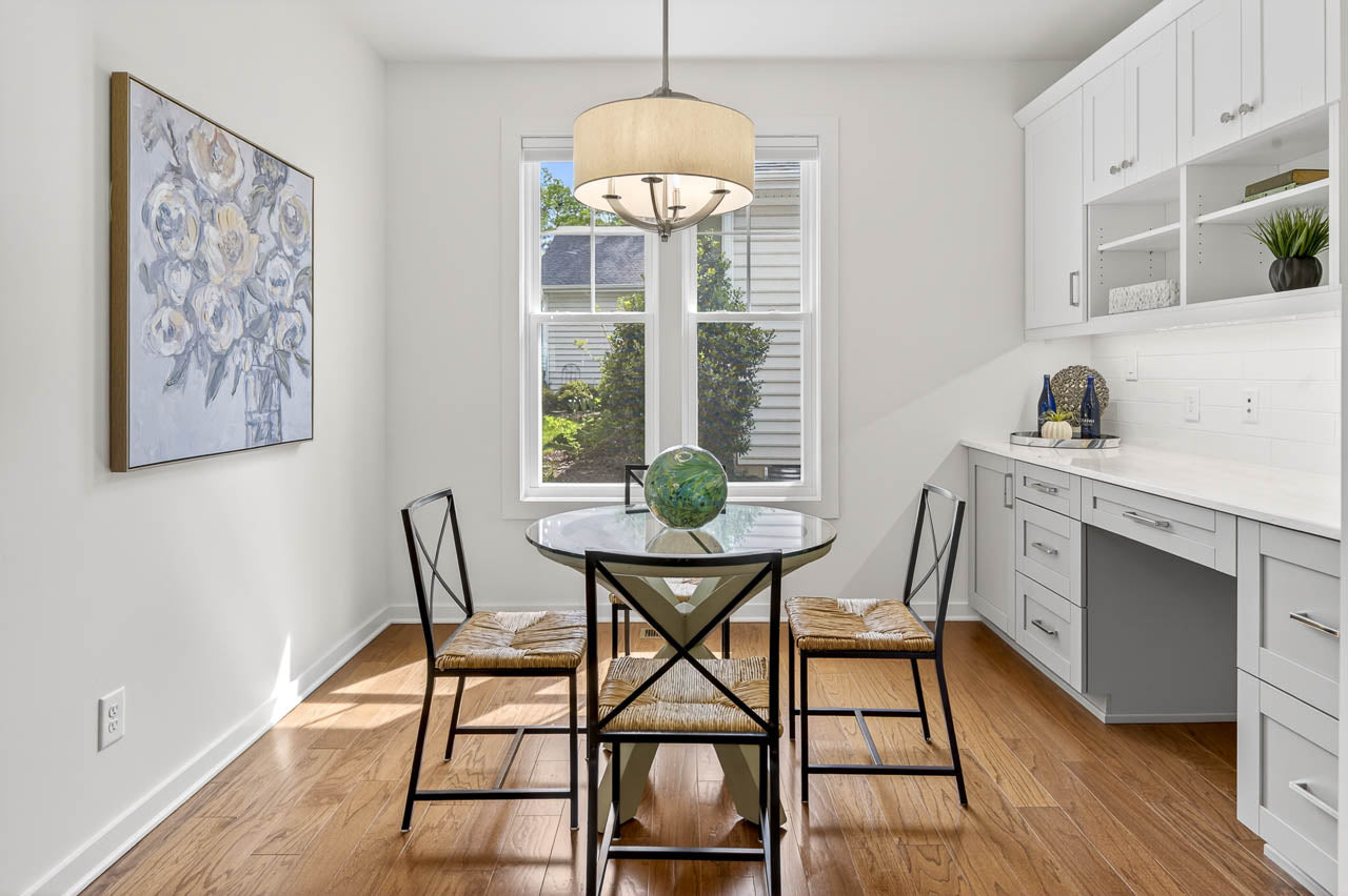 Bright dining area with a round glass table, four chairs with woven seats, a globe centerpiece, white built-in cabinets, a large floral painting, wood floor, and sunlight streaming through a window.