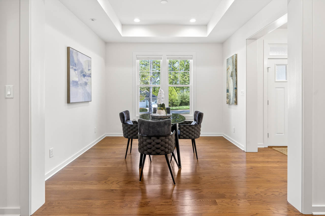 Bright dining room with wooden floors, a round glass table, and five dark upholstered chairs. Large window lets in natural light; abstract art hangs on white walls. Contemporary, minimalist design.