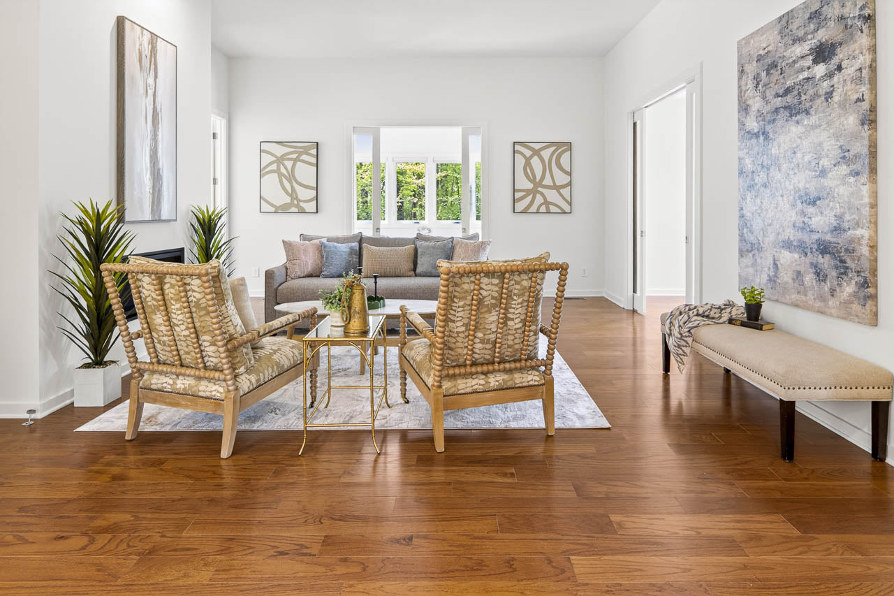 A modern living room with wooden floors, two woven chairs, a glass coffee table, a gray sofa with cushions, abstract art on white walls, green plants, and a bench near the entrance. Large windows let in natural light.