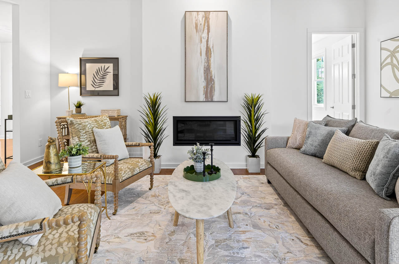 A modern living room with a gray sofa, patterned armchairs, a marble coffee table, plants, abstract wall art, and neutral decor in a bright, airy space with white walls and ample natural light.