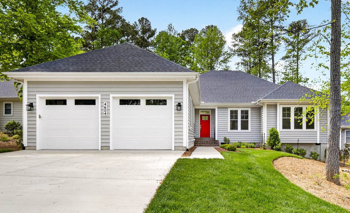 Single-story gray house with a dark shingle roof, two white garage doors, a bright red front door, front steps, and a well-manicured lawn surrounded by trees and landscaped shrubs.