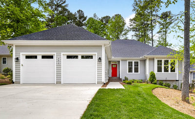 Single-story gray house with a dark shingle roof, two white garage doors, a bright red front door, front steps, and a well-manicured lawn surrounded by trees and landscaped shrubs.