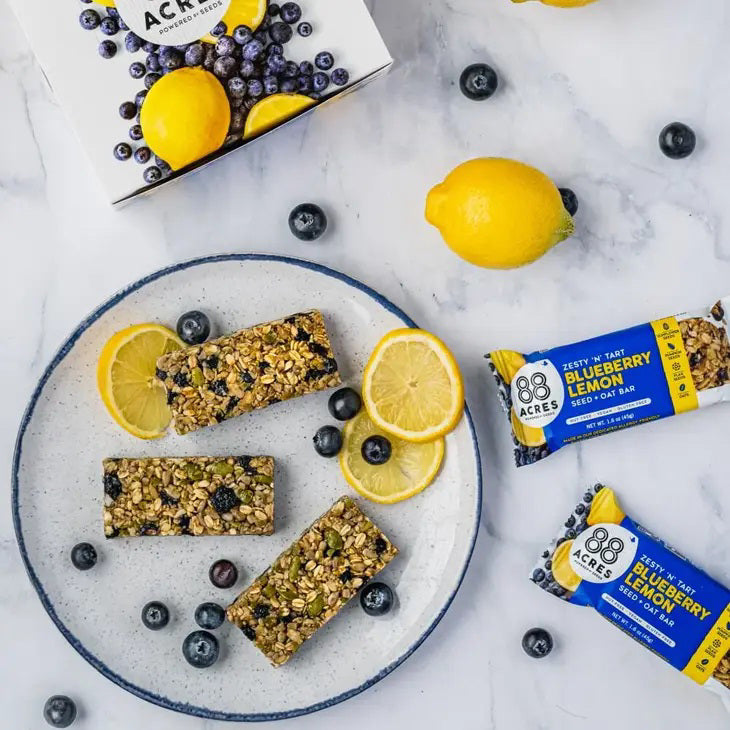 A zesty &#39;N&#39; tart 88 ACRES - BLUEBERRY LEMON SEED OAT BAR is displayed against a light wooden surface. The blue and white packaging showcases images of blueberries and lemon. This bar from 88 ACRES is nut-free, vegan, gluten-free, and made with sunflower seeds.